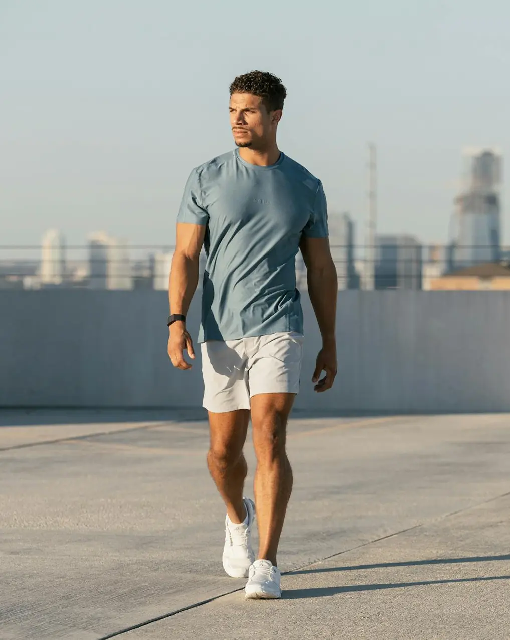 Man walking outdoors wearing a blue t-shirt, light-colored shorts, white sneakers, and a black wristband.