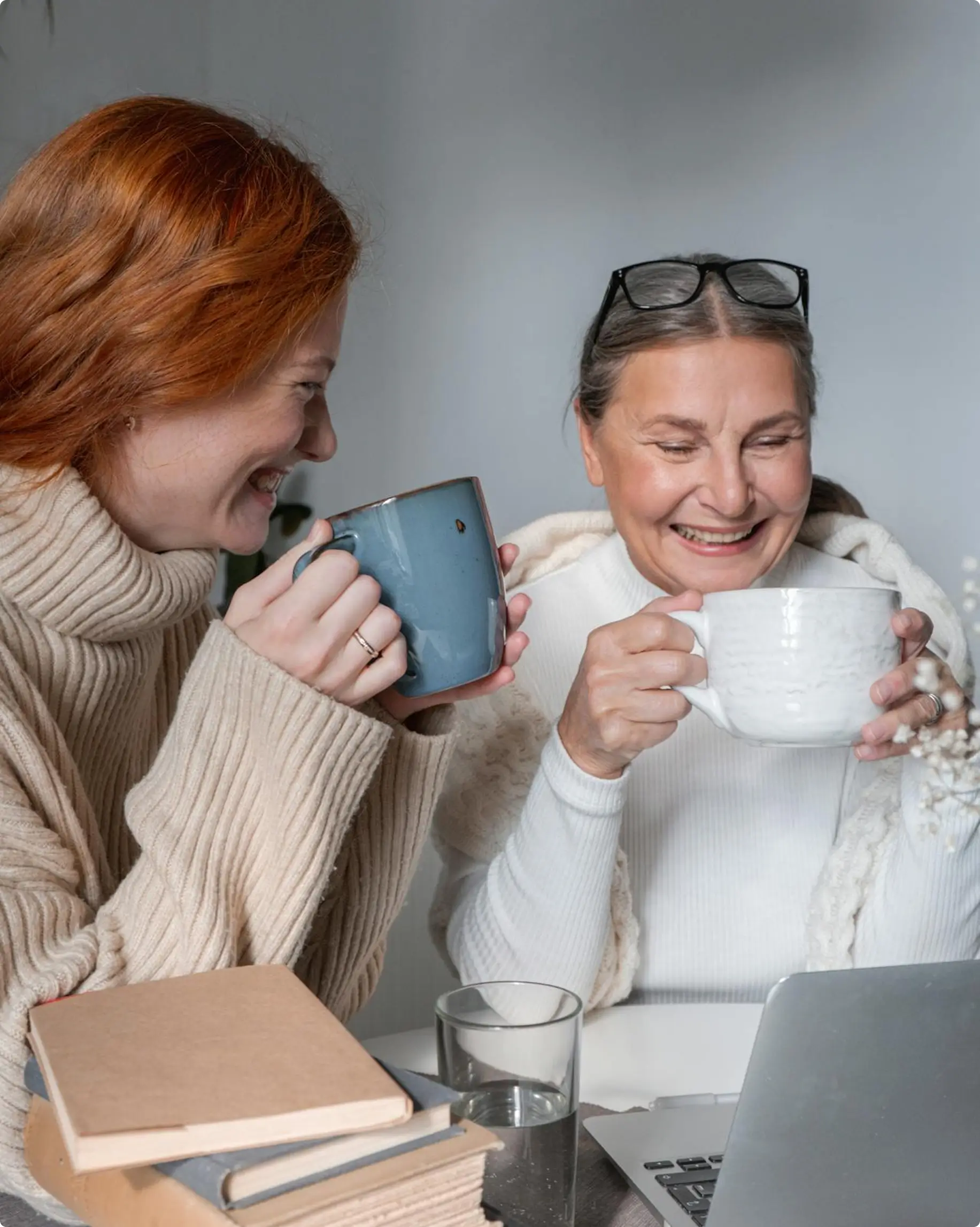 Two women smiling and holding large mugs, sitting at a table with books, a glass of water, and a laptop.